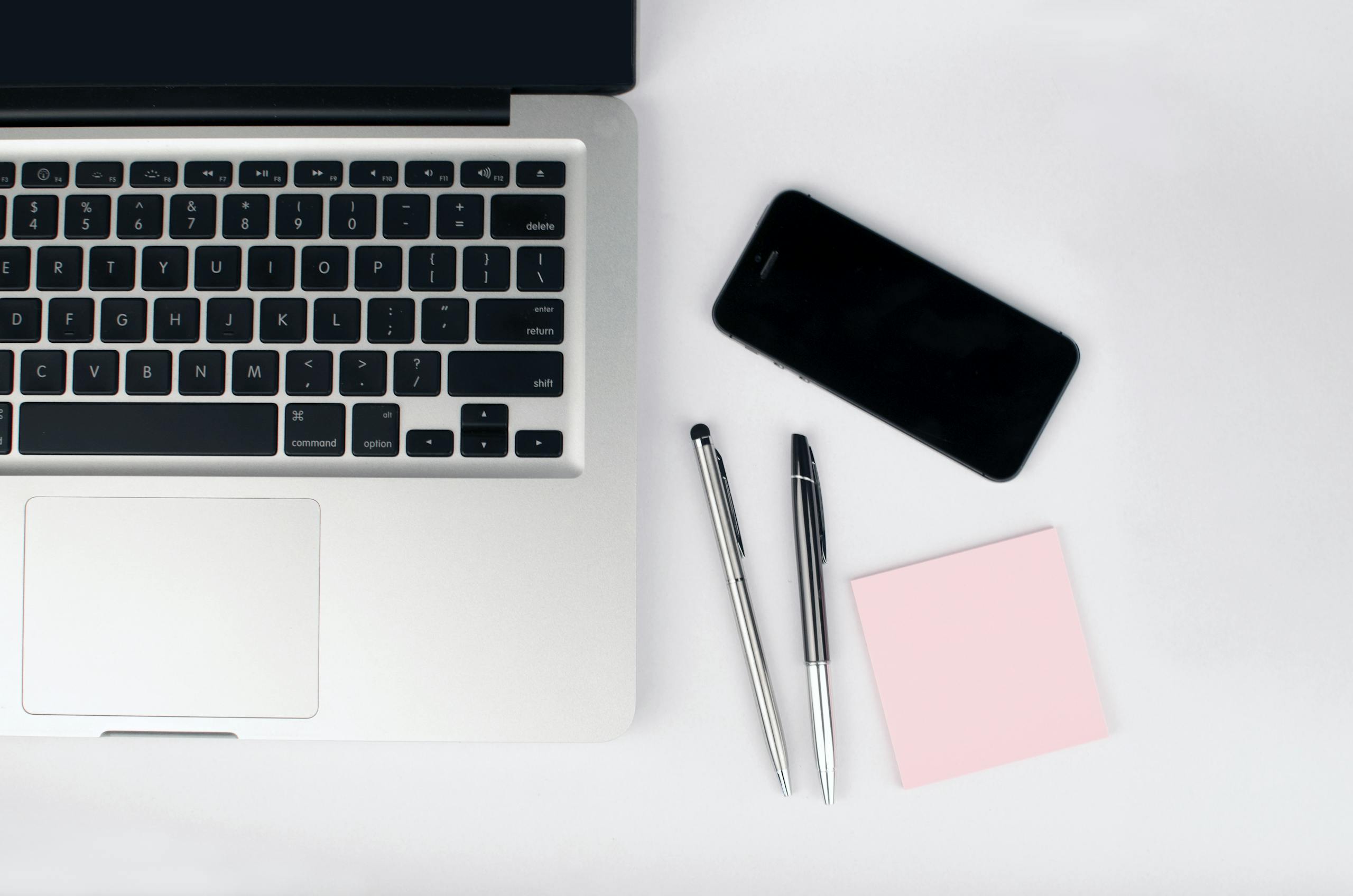 Overhead view of a sleek workspace featuring a laptop, smartphone, and stationeries on a white desk.